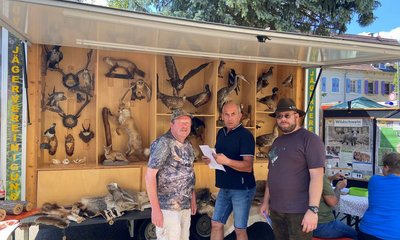 Drei Männer stehen vor einem mobilen Stand mit ausgestopften Wildtieren und Trophäen. Links und rechts sind Schilder mit der Aufschrift "JÄGERVEREINIGUNG". Im Hintergrund sind Bäume und Gebäude.